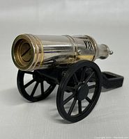 Side angle view of metal cannon lighter showing silver barrel, gold accent rings, black wheels and carriage.