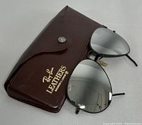 Photo showing vintage Ray-Ban aviator sunglasses with reflective mirrored lenses positioned in front of original brown leather case stamped 'Ray-Ban LEATHERS Sunglasses 100% UV'