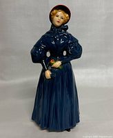Front view of the porcelain figurine showing the woman's face, bonnet, and blue dress with a small bouquet in her hands.