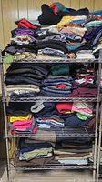 Metal shelving unit with multiple shelves holding folded clothing in assorted styles, sizes, and colors. Clothes organized in stacks but not further detailed.
