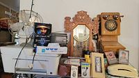 Wide view of lot showing vintage wooden phone, ornate clock, electric fan, boxed electronics and accessories