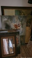 Stack of multiple framed paintings leaning against a wall on a floor showing partially visible art including a figure scene and landscapes.