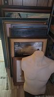 Framed pictures stacked against the wall in various styles and sizes with a beige male torso mannequin standing in front