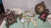 Photo showing an assortment of Victorian-style ornate metal photo frames with vintage photos, a floral-patterned trinket box, vanity items including a hand mirror, hairbrush, small tray, a glass perfume bottle, and a braided decorative headband or belt