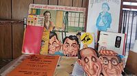Display of vintage advertising posters, papers, and three exaggerated caricature masks arranged on a table and propped against a wooden wall.