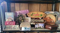 Shelf display with variety of vintage and modern decorative boxes, trays, and signs including floral boxes, painted trays, and small ceramic pieces.