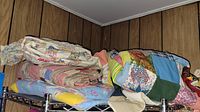 Stack of folded vintage quilts and blankets on metal shelving against a paneled wall.