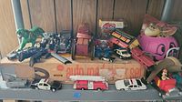 Overview of assorted vintage toys including MOTU action figures, slot cars, diecast fire trucks, police vehicles, a green tiger figure, and wooden fire engine toy on a box.