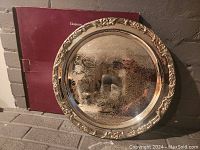 Front view of the 17-inch round electroplated steel tray leaning against the burgundy original box on a brick surface. Shows the embossed decorative border and fine engraved pattern on tray surface.