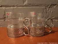 Two clear Iittala Tsaikka glass coffee/tea cups with distinctive nickel-plated mesh holders and curled metal handles sitting side by side, showing full profile and handles.