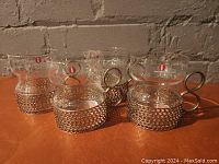 Four clear glass coffee/tea cups with hourglass shape, each sleeved in nickel-plated mesh metal holders with circular handles, positioned on an orange surface against a textured brick background.