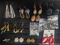 Full view of 14 pairs of costume earrings on black background showing different styles including long silver dangles, iridescent and shell style, tassels, and clip-ons.