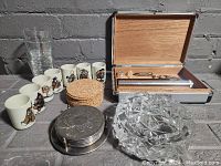 Full lot showing humidor box with accessories open, set of 7 saki cups decorated with dog motifs, several cork coasters stacked alongside, embossed metal coaster holder, and cut glass ashtray on a gray surface.