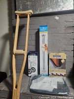 Photo showing the lot items: one wooden crutch, boxed adjustable shower grab bar, boxed MaxTid door draft stopper, and a laundry bag with wheels.