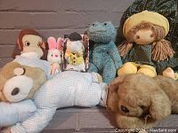 Seven different stuffed animals and dolls including rag doll, bear, dog, monkey, bunny, mouse, and frog arranged on the ground for listing.
