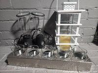 Lot arranged with white wooden outdoor lantern on right, two black LED water lights with glass tops in center, and five clear glass tea light holders in front on a hammered metal tray.