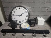 Photo showing round wall clock, black wire mesh pencil holder, small wooden cube, round tape container, black 5-hook rack on table against brick wall.