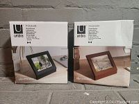 Boxes of two Umbra Podium photo frames, one black and one brown wood finish, both showing front and side images and packaging with branding and size details.