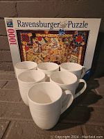 Puzzle box with colorful map design showing completed puzzle image behind six white ceramic mugs.