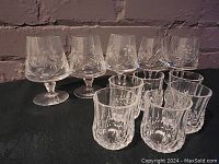 Set of 5 cocktail glasses with etched cornflower floral pattern and 6 clear textured shot glasses on a surface against a brick wall.