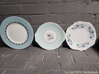 Group shot of three decorative porcelain plates showing a pale blue rim plate, a pale blue handled plate with floral center, and a white scalloped plate with blue flowers.