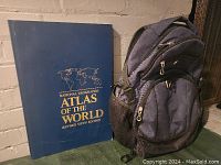 Photo showing the blue cover of the 1992 National Geographic Atlas of the World and the dark blue Swiss Gear backpack side by side.