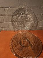 Photo shows two glass serving platters against a brick wall background on an orange surface, one with floral embossed pattern and scalloped edges and the other with ribbed swirl design.