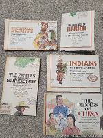 Five National Geographic supplemental booklets laid out on carpet, covers visible with titles and illustrations representing African, Pacific, Southeast Asian, North American Indian, and Chinese cultures.