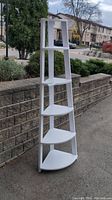 Front and side views of a white wooden corner book shelf with five triangular shelves outdoors by a stone wall.