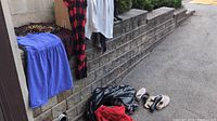 Variety of women's clothes including blue skirt/pants, red and black plaid shirt or dress and white and black tops hung on a low retaining wall with some clothes in black plastic bags on the ground.
