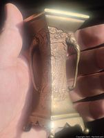 Photo of small bronze vase held in hand showing square tapered body, detailed engravings, and elephant head handle with trunk extending down the side.