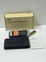 Digital moisture meter device with LCD screen and orange buttons, laid beside original packaging box, two AA batteries, and user manual on a white surface.