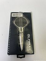 Front view of Global kitchen shears in black box with transparent window showing the shears, minor damage to box edges visible.