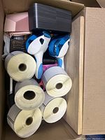 Opened cardboard box showing five large rolls of adhesive labels or receipt tape, two handheld garment steamers, and smaller boxed items underneath.