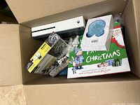 Overview of box showing white Xbox One S console, collectibles, blue clock in box, Christmas decor, and model vehicle