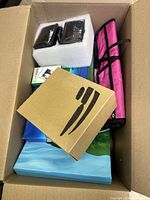 Top-down view inside box showing two small rectangular black lights or lenses in foam, several boxed items, and pink zippered case.