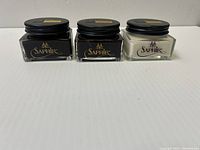 Three jars of Saphir Medaille d'Or Pommadier Cream in black, brown, and neutral colors placed side by side on a white surface.