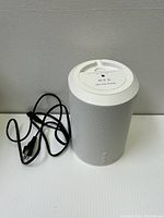 White Sonos Era 100 cylindrical wireless speaker standing on a white surface with black power cord beside it.