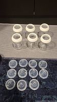 Six clear Tommee Tippee glass baby bottles with white screw-on rings arranged in two rows on a grey textured surface; below are 11 clear silicone nipples arranged in rows on a dark blue surface.
