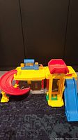 Yellow and red Fisher-Price Little People playset with a blue slide and a red curved ramp, placed on a dark floor with a black background.