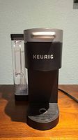 Front view of black and silver Keurig Supreme coffeemaker on wooden surface with 'Keurig' branding visible on front.