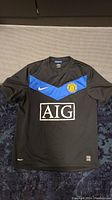 Black Manchester United soccer jersey laid flat, showing blue chevron across chest with AIG logo and club crest.