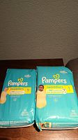 Photo shows two Pampers Swaddlers diaper packages on a dark table with a textured wall in the background. One pack is size NB with 31 diapers and the other is size 1 with 32 diapers, both in turquoise packaging.