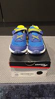 Front facing blue and yellow Saucony kids shoes placed on the original box showing shoe design and details.