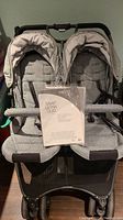 Gray Valco Baby Snap Ultra Duo double stroller with manual displayed on one seat, showing dual seats, canopies, safety harnesses, and padded safety bars.