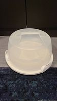 Round white cake tray with clear dome cover and integrated handle on top, sitting closed on a blue surface.
