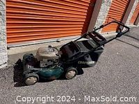 Side view of green Craftsman lawn mower showing beige engine cover and left wheel on pavement outside storage units.