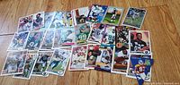 Photo showing over 30 football trading cards spread on a wooden floor. Cards include various teams such as Jets, Orioles, Raiders, Cowboys, Rams among others. Cards show players in action poses and close-up portrait styles.