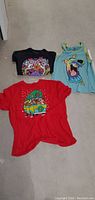 Three Scooby Doo tops laid out on the floor: red T-shirt, black T-shirt, and blue tank top
