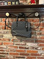 Black Nine West purse hanging on a rustic brick wall with front view showing the bag's shape, handles, shoulder strap, and charm.
